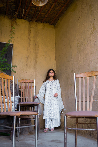 Woman in a white traditional outfit standing in a rustic outdoor setting with wooden chairs.