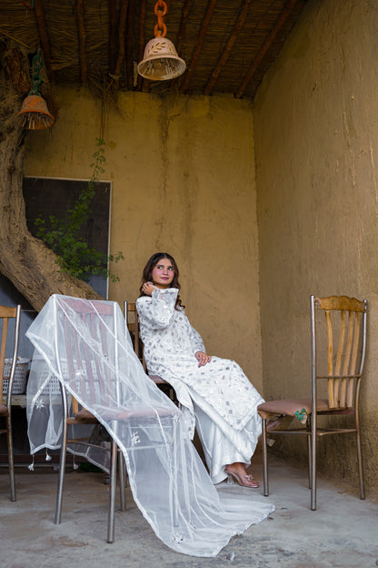 Woman in a white outfit sitting on a chair with a sheer white fabric draped over it in a rustic setting.